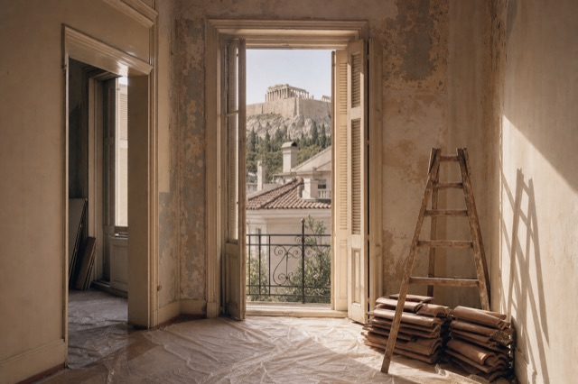 Sunlight falling through tall windows into an Athens apartment under renovation, dust sheets on the floor, the Acropolis visible outside