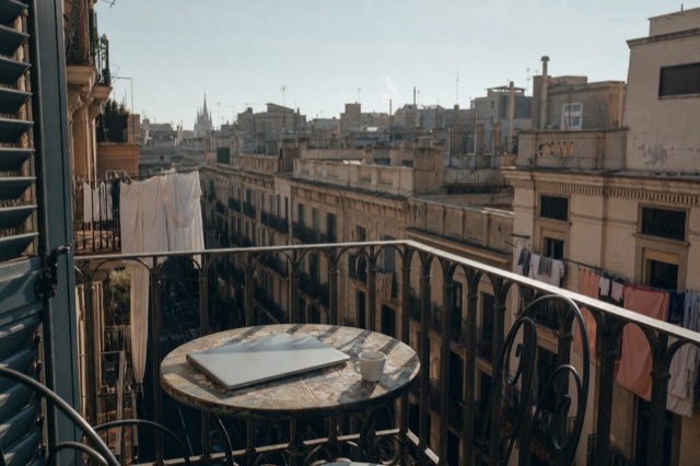 A closed laptop and an espresso cup on a wrought-iron balcony table above Barcelona rooftops in morning light