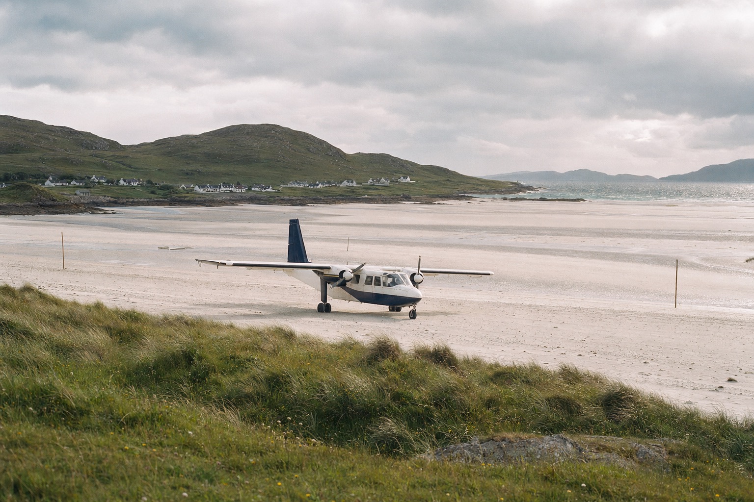 A Britten-Norman Islander aircraft on a wide white-sand beach at low tide on the Isle of Barra, with the green hills and Castlebay village in the distance, soft northern light