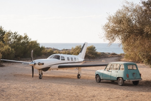 A small white Piper Meridian on a sandy airstrip in the Balearics, late afternoon light, sea visible behind low scrubby pines