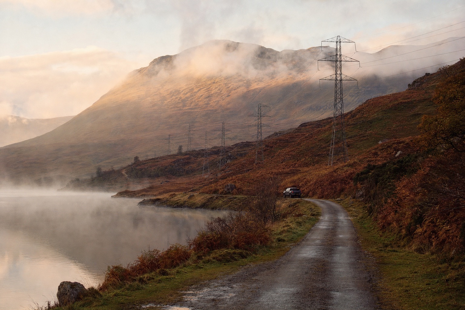 A single-track Highland road running beside a loch toward a steel pylon line under a low morning sky