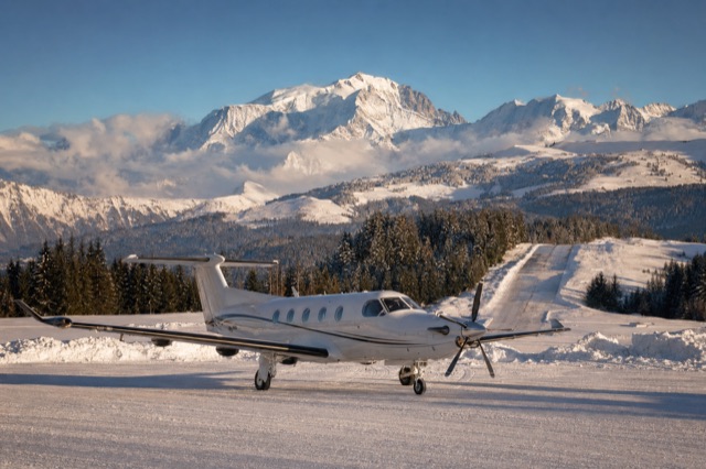 A Pilatus PC-12 aircraft on the snow-covered apron at Megève altiport with the Mont Blanc massif in the background, blue sky, morning light