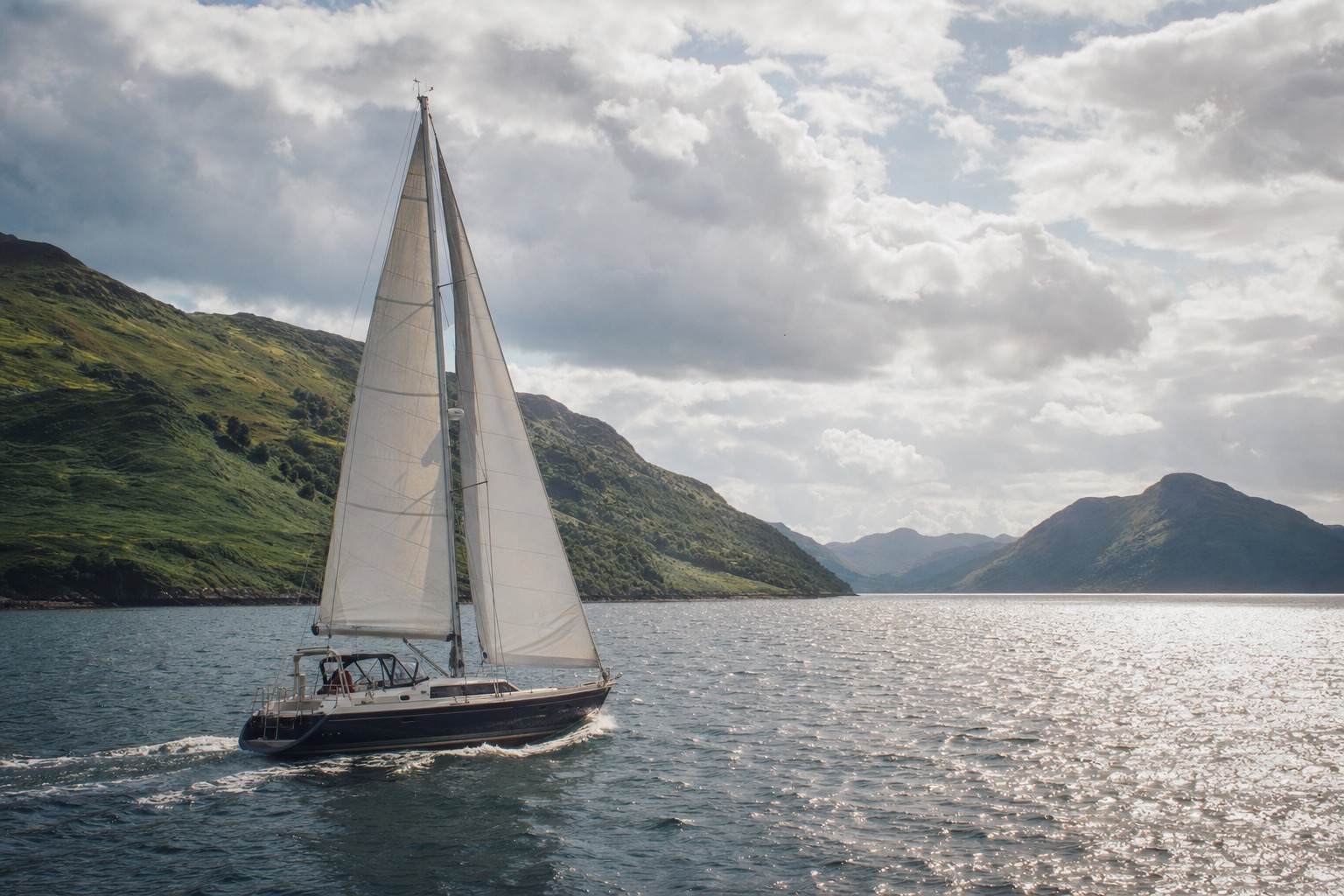 A cruising yacht under sail in the Sound of Mull on a bright morning, green hills of Mull on one side and the Morvern shore on the other